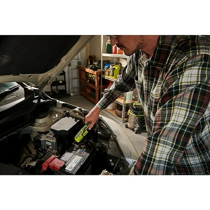 Ryobi 4V USB Akku-Ratschenschrauber RR14W4-0 - 4 V, Ohne Akku - Ein Mann, der mit einer gelben Ryobi-Lampe unter der Motorhaube eines Autos hantiert, wobei der Fokus auf der Lampe und der Autobatterie liegt.