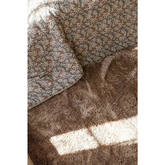 Manta para sofá Otoño - Textil, Azul/Crudo - Primer plano de un elegante cojín con estampado de hojas sobre una alfombra marrón, iluminado por un brillante rayo de sol.