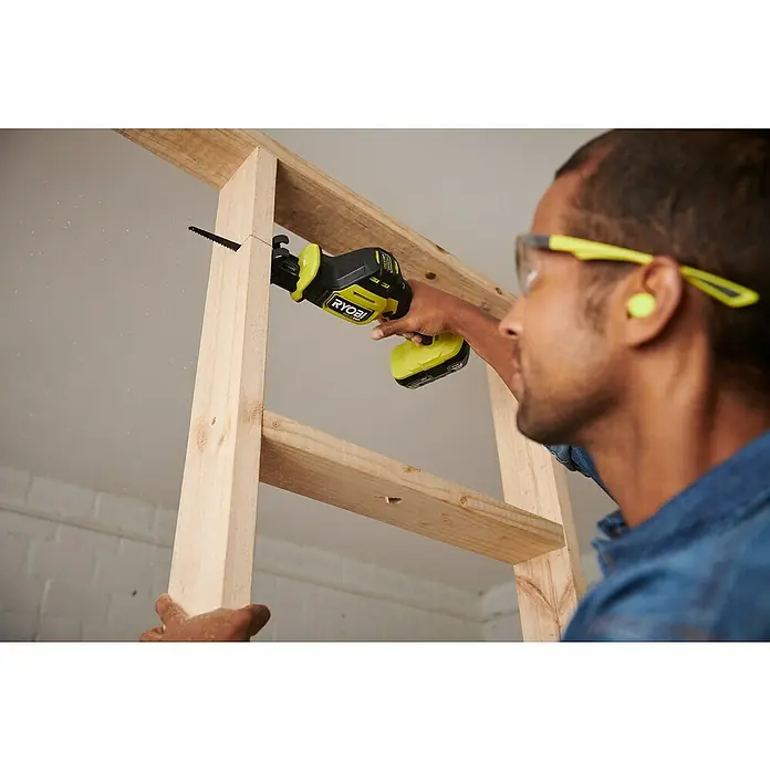 Ryobi ONE+HP Akku-Säbelsäge RRS18C1-0 - 18 V, Leerlaufdrehzahl: 0 U/min - 3.200 U/min, Hublänge: 23 mm - Nahaufnahme einer Person, die mit einer gelb-schwarzen Ryobi Säbelsäge ein Holzstück durchsägt. Sie trägt Schutzbrille und Ohrstöpsel.