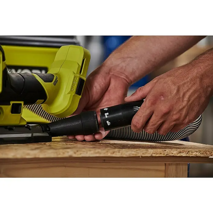 Ryobi ONE+HP Akku-Pendelhubstichsäge RJS18BX-0 - 18 V, Li-Ionen, Anzahl Akkus: Ohne Akku, Leerlaufhubzahl: 900 /min - 3.200 /min - Ein Mann verbindet einen Staubsaugerschlauch mit einem gelben Ryobi ONE+ Akku-Bandschleifer auf einer Holzoberfläche.