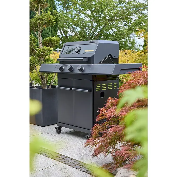 Kingstone KMS Gasgrill Cliff 3000 Beast Black Edition - Grillfläche (B x T): 61 x 45 cm, 10,5 kW, Anzahl Brenner: 3 Stk. - Schwarzer Grill mit Bedienknöpfen und Schrank. Der Grill steht auf einer Terrasse und ist von Pflanzen umgeben.