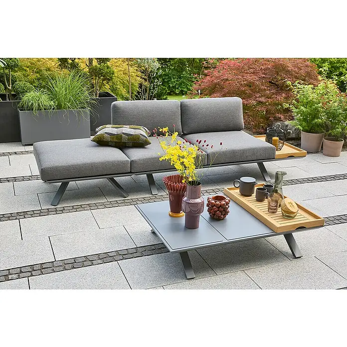 Sunfun Loungemöbel-Set Lumar - 4 -tlg., Aluminium, Teak, Ablagefläche links, Night-Grey - Graue Outdoor-Lounge-Garnitur mit Couch und Tisch auf einer Terrasse, umgeben von Pflanzen und Bäumen. Perfekt zum Entspannen im Freien.