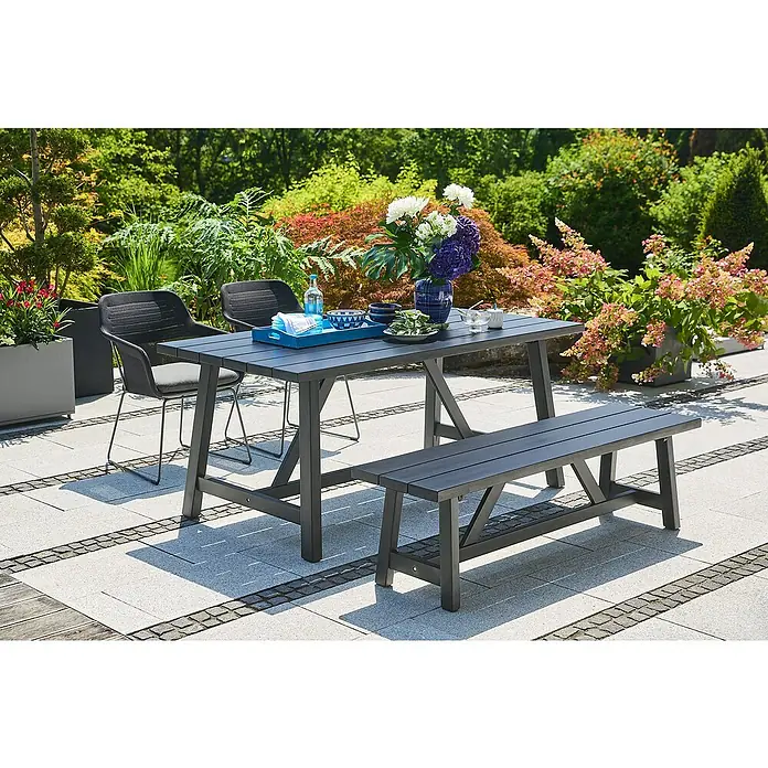 Sunfun Gartenmöbel-Set Melina - 4 -tlg., Akazie/Aluminium/Polyester, Black Washed/Night-Grey - Grauer Gartentisch mit passender Bank und zwei Stühlen auf einer gepflasterten Terrasse, umgeben von üppigem Grün.
