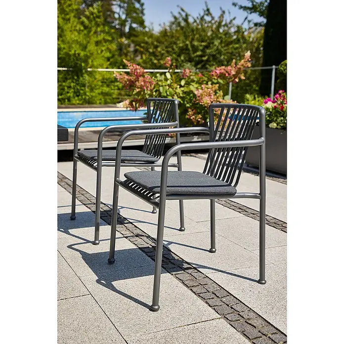 Sunfun Sarah Stapelsessel - Stapelbar, Night-Grey, B x T x H: 59 x 80 x 57 cm - Zwei graue Armlehnstühle mit Kissen stehen auf einer Terrasse mit Pool im Hintergrund.