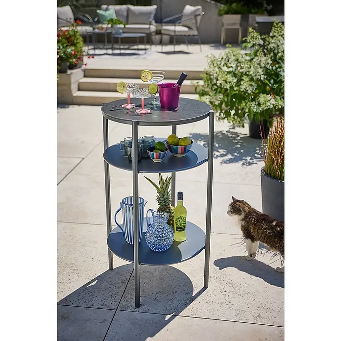 Sunfun Solo Garten-Beistelltisch rund - Ø x H: 65 x 108 cm, Stahl, Night-Grey - Ein grauer, dreistöckiger Servierwagen steht im Freien, beladen mit Getränken, Obst und Eis. Eine Katze steht daneben.