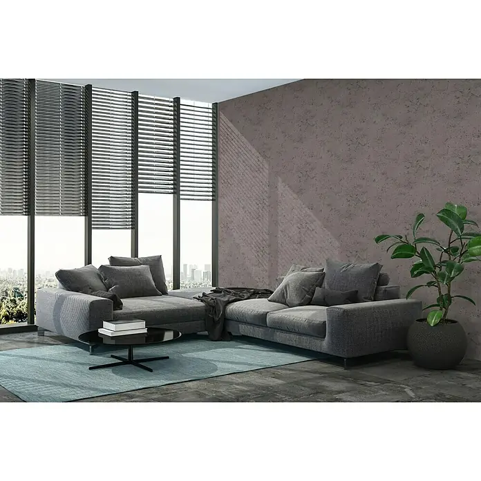 A.S. Création New Walls Vliestapete Used Wall - Anthrazit, Betonoptik, 10,05 x 0,53 m - Graues Ecksofa mit Zierkissen und einem Couchtisch auf einem blauen Teppich, neben einem Fenster mit Jalousien und einer Zimmerpflanze.