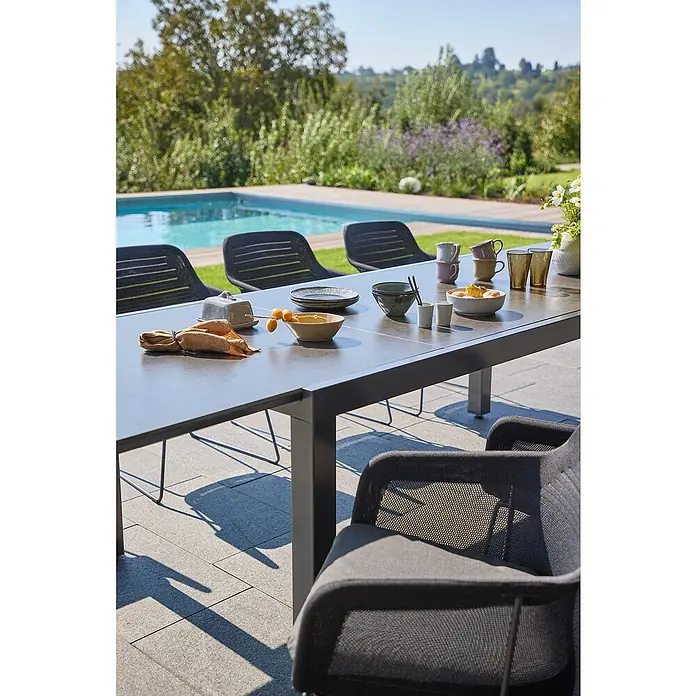 Sunfun Gartenmöbel-Set Maja - 9 -tlg., Night-Grey, Ausziehbar, Tisch (L x B x H): 200-300 x 100 x 74 cm, Stuhl (B x T x H): 57 x 65 x 83 cm - Ein moderner Esstisch aus grauem Metall mit Geschirr im Freien neben einem Pool, der sich ideal für Mahlzeiten im Freien eignet.