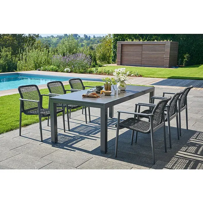 Sunfun Gartenmöbel-Set Maja/Solo - 7 -tlg., Night-Grey, Ausziehbar - Grauer Gartentisch mit sechs Stühlen aus Korbgeflecht auf einer Steinterrasse neben einem Pool. Im Hintergrund ist ein Gartenhaus sichtbar.
