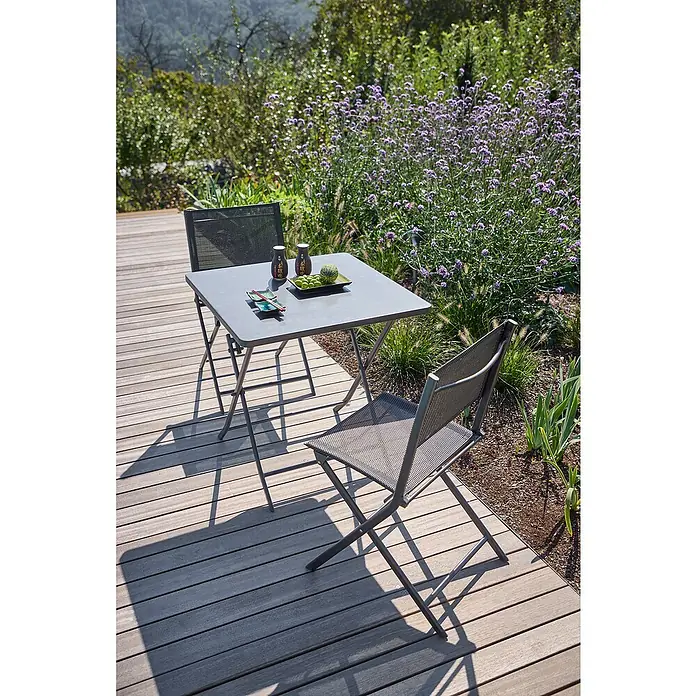 Sunfun Balkonmöbel-Set Maja - 3 -tlg., Night-Grey, Tisch (LxBxH): 70 x 70 x 71 cm, Stuhl (TxBxH): 46 x 52 x 81 cm - Grauer Gartentisch mit zwei Stühlen auf einer Holzterrasse, gedeckt mit asiatischem Geschirr, umgeben von üppiger Blumenpracht.