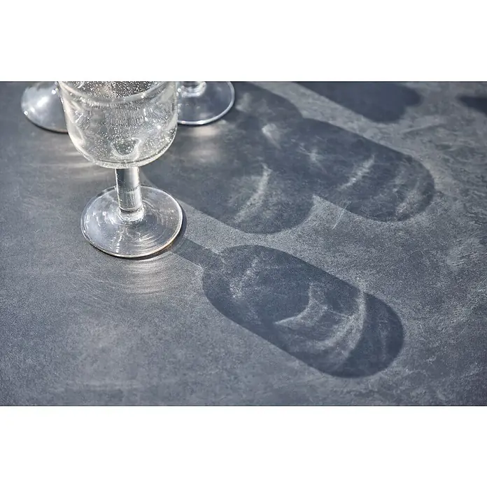 MFG Gartentisch Boulevard - L x B: 110 x 70 cm, Stahl, Mecalit, Schiefer, Klappbar - Ein schattiges Arrangement zeigt elegante, leere Cocktailgläser auf einer dunklen Oberfläche, die ein interessantes Lichtspiel erzeugt.