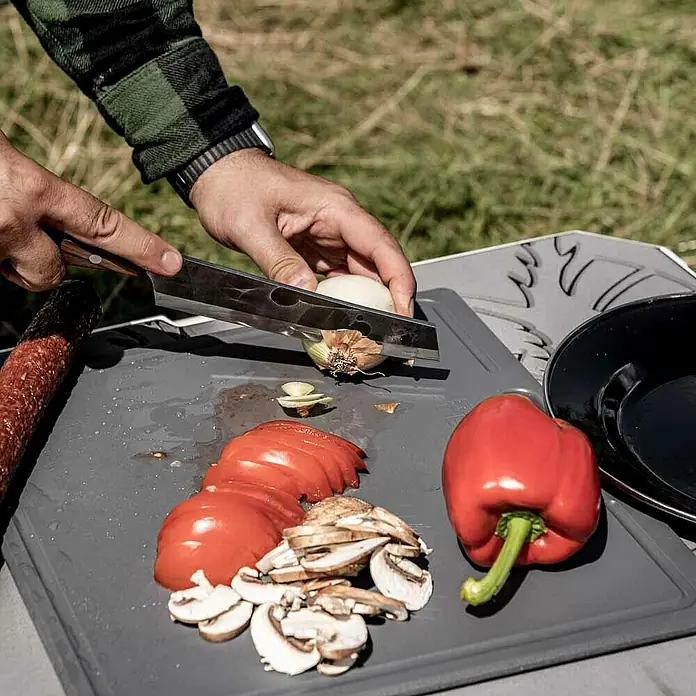 Petromax Schneidebrett - Passend für: Petromax Kühlbox 25l, Kunststoff - Ein Mann schneidet eine Zwiebel auf einem Schneidebrett mit Gemüse, darunter Paprika, Tomaten und Pilze, im Freien.