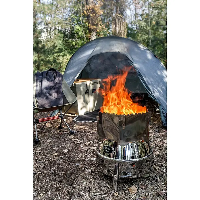 Petromax Grillaufsatz Atago - Passend für: Petromax Grillserie Atago, Edelstahl - Nahaufnahme eines Petromax Atago Grills mit hellem Feuer, umgeben von Campingausrüstung wie Zelt und Stuhl.