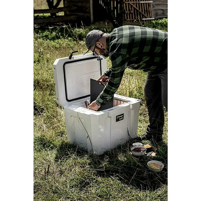Petromax Schneidebrett - Passend für: Petromax Kühlbox 25l, Kunststoff - Eine Person beugt sich zu einer Kühlbox der Marke Pelican, die im Freien auf Gras steht, daneben Schalen mit Lebensmitteln.