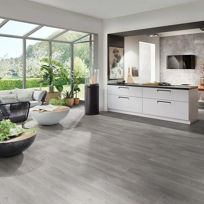b!design Vinylbodenmuster Rigid SPC Swiss Pinie - 281 x 161 x 3,8 mm, Landhausdiele - Einladender Wohnraum mit modernem Küchenbereich und stilvollem grauem Holzboden, der Wärme und Eleganz ausstrahlt.