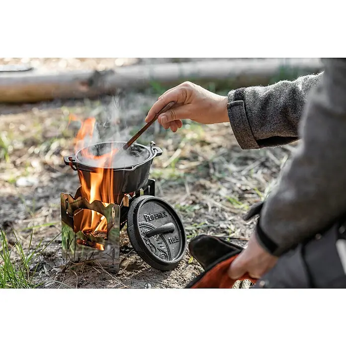Petromax Campingkocher Hobo - B x T x H: 14 x 11,7 x 12,5 cm, Edelstahl - Ein Feuertopf steht auf einem Hobo-Kocher im Freien, während eine Hand mit einem Löffel den Inhalt umrührt. Der Deckel liegt daneben.