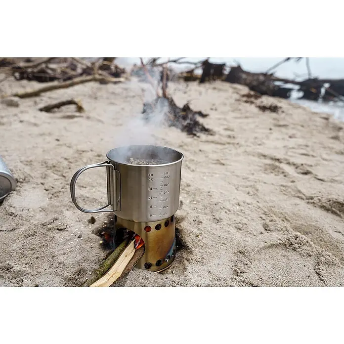 Petromax Kochbecher - Edelstahl - Edelstahlbecher erhitzt auf einem Campingkocher am Strand. Der Becher steht auf einem Lochkocher mit brennenden Holzscheiten darin.