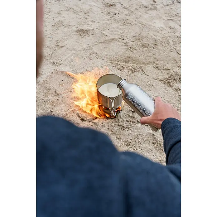 Petromax Kochbecher - Edelstahl - Eine Person gießt Flüssigkeit aus einer gemusterten Metallflasche in einen Topf, der über einer offenen Flamme steht.