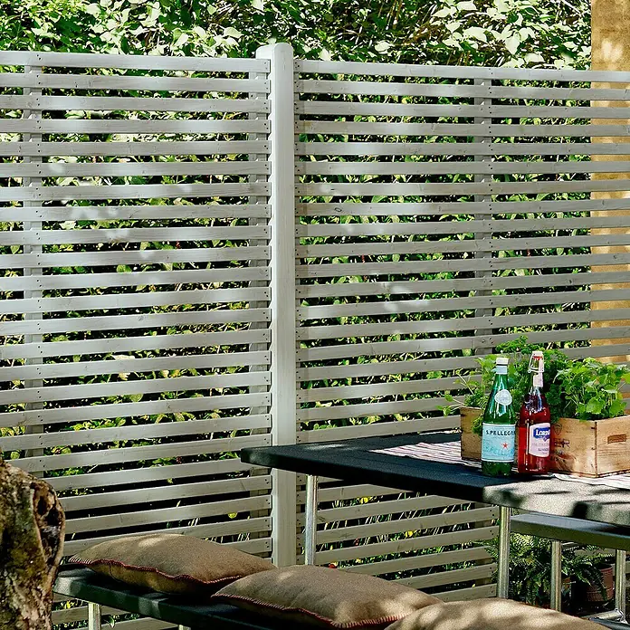 Plus Sichtschutzelement Tokyo - 180 x 180 cm, Holz, Weiß RAL 9010 - Ein Außenbereich mit einem Holzzaun, einem Tisch mit Getränken und einem Sitzbereich mit Kissen, der eine entspannende Atmosphäre bietet.