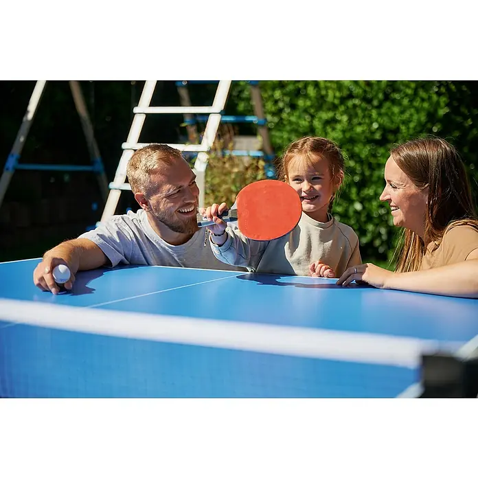 Hudora Tischtennisplatte Match - Blau - Familie spielt Tischtennis im Freien. Der Fokus liegt auf dem blauen Tischtennistisch mit Netz, Schläger und Ball, umgeben von lächelnden Personen.