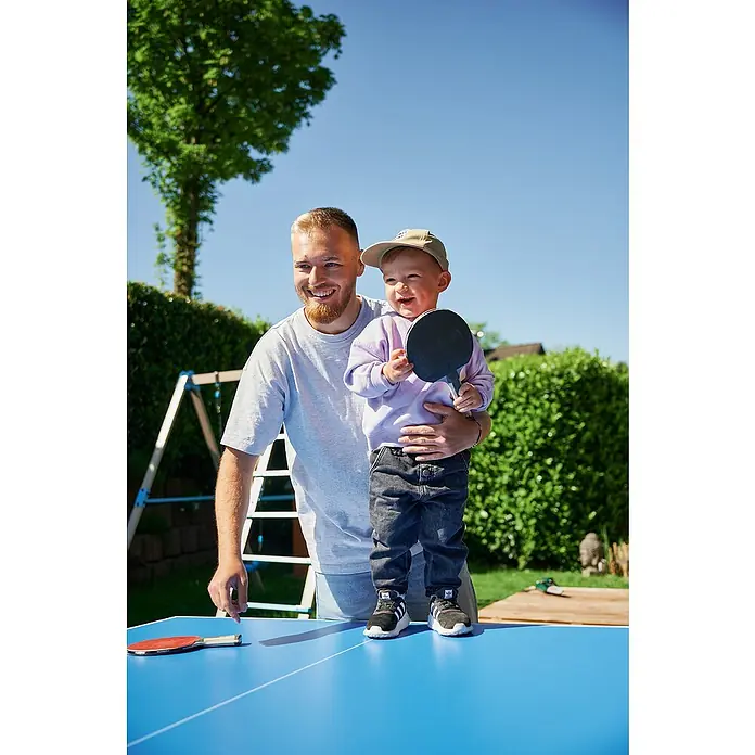 Hudora Tischtennisplatte Match - Blau - Ein Vater hält seinen Sohn auf einem Tischtennistisch. Beide haben Spaß und halten Tischtennisschläger in der Hand.