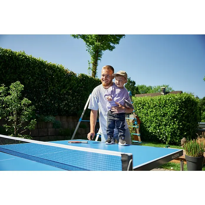 Hudora Tischtennisplatte Match - Blau - Ein Vater hält sein Kind auf einem blauen Hudora Tischtennistisch im Freien, mit Schlägern bereit für eine Partie.