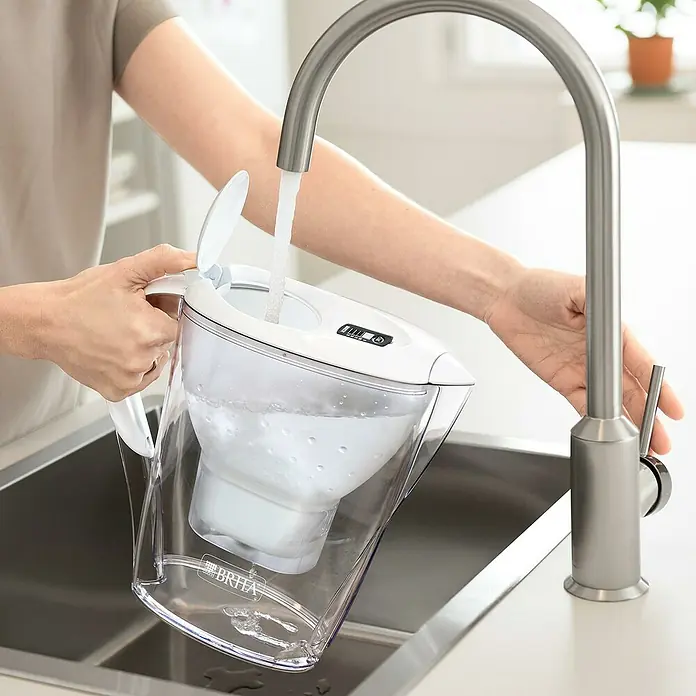 Brita Jarra filtrante Marella - Capacidad volumétrica: 2,4 l, 1 ud., Transparente/Blanco - Una persona llena una jarra de filtro de agua Brita debajo de un grifo, filtrando y purificando el agua del grifo.