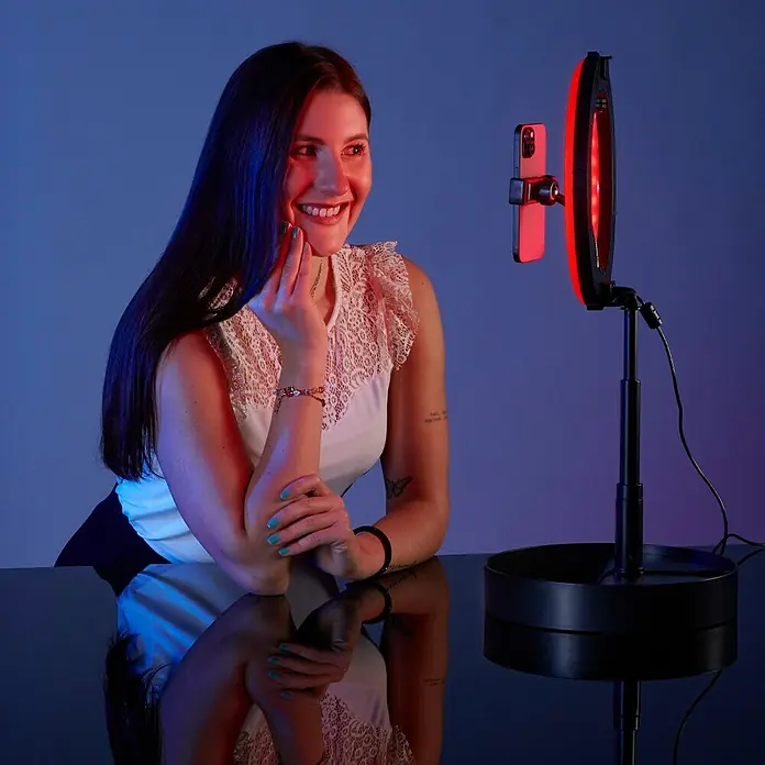 Hama Smartphone-Halter LED-RGB-Ringleuchte "SpotLight FoldUp RGB 102" - Schwarz - Die Frau benutzt eine Studioleuchte mit Smartphone-Halterung, um professionelle Inhalte zu erstellen oder an Videoanrufen teilzunehmen.