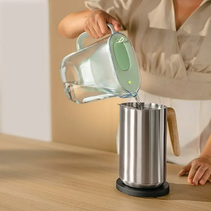 Brita Cartucho para jarra filtrante Maxtra Pro Experto en Cal - Específico para: Jarras filtrantes Brita, 4 pzs. - Mujer vertiendo agua de una jarra de filtro de agua Brita en un elegante recipiente de acero inoxidable con mango de madera.