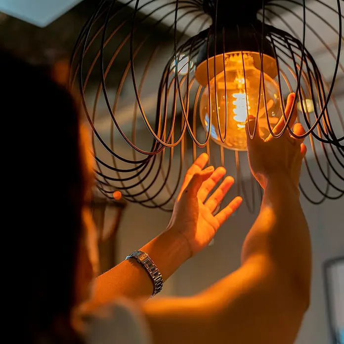 Hama Wifi Smart-LED-Lampe Filament Matter - E27, Dimmbar, RGBW, 470 lm, 4,9 W - Eine Nahaufnahme einer Glühbirne in einer dekorativen, schwarzen Drahtleuchte, die von zwei Händen berührt wird.
