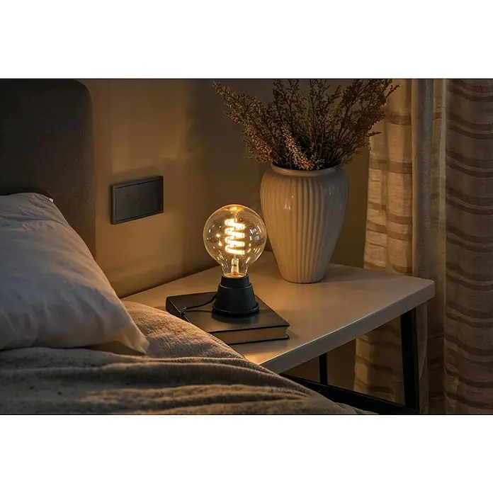 Hama Wifi Smart-LED-Lampe Filament Matter - E27, Dimmbar, RGBW, 470 lm, 4,9 W - Nachttischlampe im Schlafzimmer. Die Glühbirne gibt ein warmes Licht. Eine Vase mit Blumen und ein Buch vervollständigen das Ambiente.