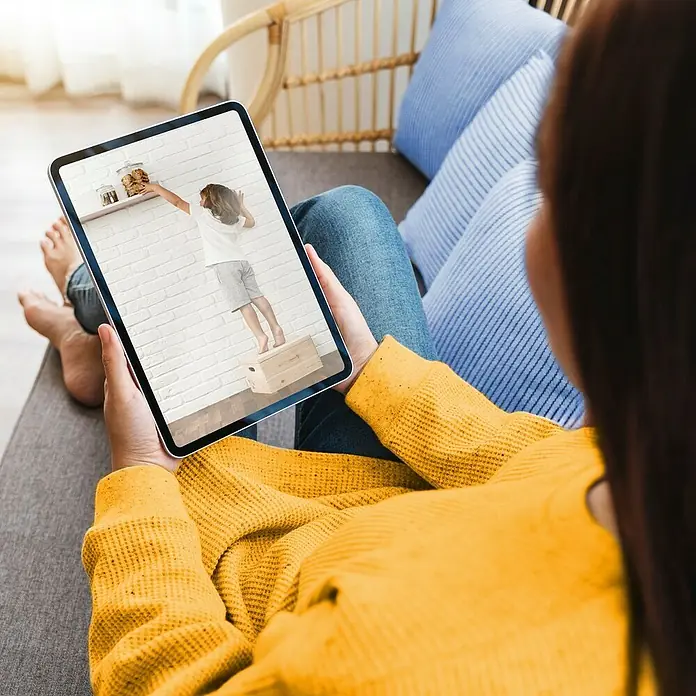 Hama Wifi Überwachungskamera - Erfassungswinkel: 100 ° - Frau entspannt auf Couch und schaut auf einem Tablet ein Video von einem Kind, das auf einen Hocker steigt, um Kekse zu erreichen.