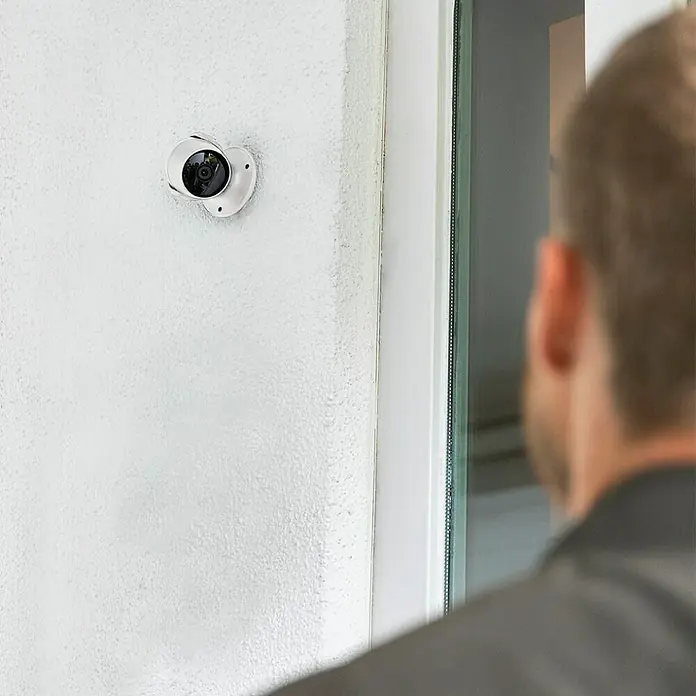 Hama Wifi Überwachungskamera 176645 - Erfassungswinkel: 110 ° - Eine Nahaufnahme einer weißen Überwachungskamera, die an einer Wand montiert ist, mit einer Person, die im Hintergrund steht.