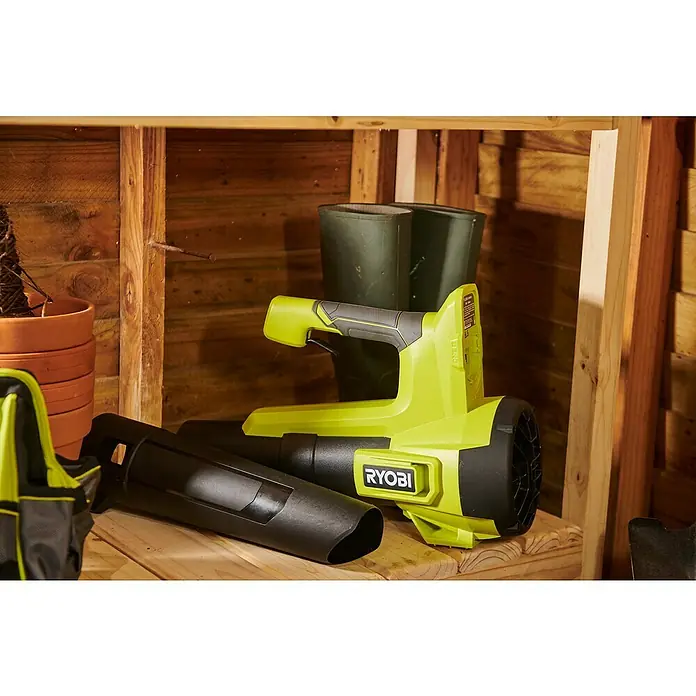 Ryobi ONE+ Akku-Gebläse RY18BLB-0 - 18 V, Ohne Akku, Blasgeschwindigkeit: 160 km/h - Ein Ryobi Laubbläser in Grün und Schwarz liegt auf einem hölzernen Regal, komplett mit Aufsatz, neben grünen Gummistiefeln.
