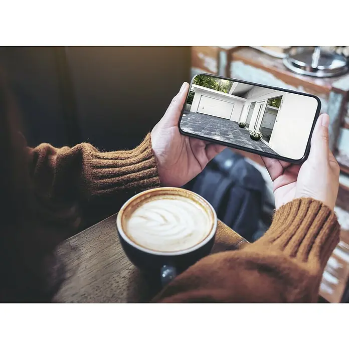 Hama Wifi Überwachungskamera 176645 - Erfassungswinkel: 110 ° - Die Person hält ein Smartphone und betrachtet ein Garagentor-Foto, während sie einen Cappuccino auf einem Holztisch genießt.
