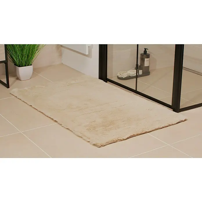 Badteppich Happy - 67 x 110 cm, Beige, 100% Polyester - Weicher beiger Badezimmerteppich liegt auf hellen Fliesen neben einer Duschkabine und einer Pflanze, der für Komfort und Stil im Badezimmer sorgt.