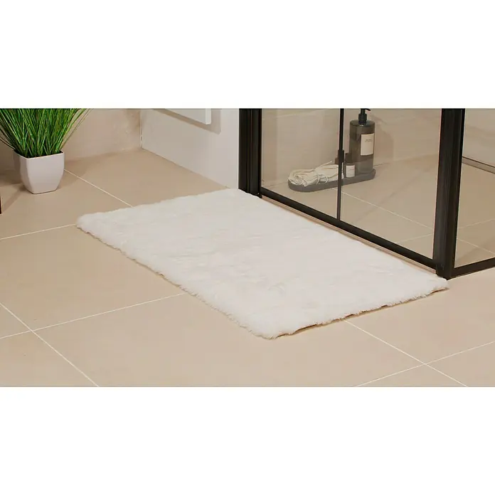 Badteppich Happy - 50 x 90 cm, Weiß, 100% Polyester - Ein rechteckiger, weicher, weißer Badteppich liegt auf hellen Fliesen vor einer Duschkabine, neben einer Topfpflanze, für ein luxuriöses Badezimmerambiente.