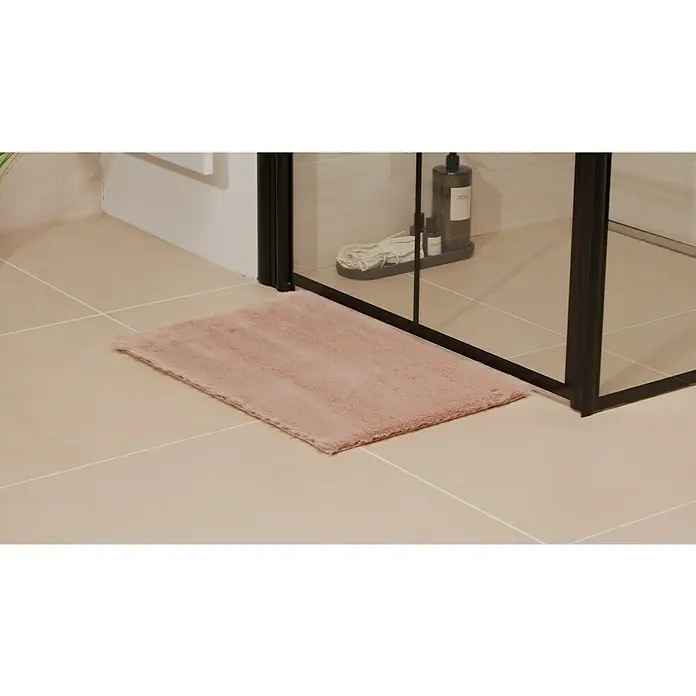 Badteppich Happy - 40 x 60 cm, Rosa, 100% Polyester - Rosa Badematte vor einer Duschkabine auf beigefarbenen Fliesen. Die Matte ist flauschig und rechteckig. Badezimmer-Accessoires im Hintergrund.