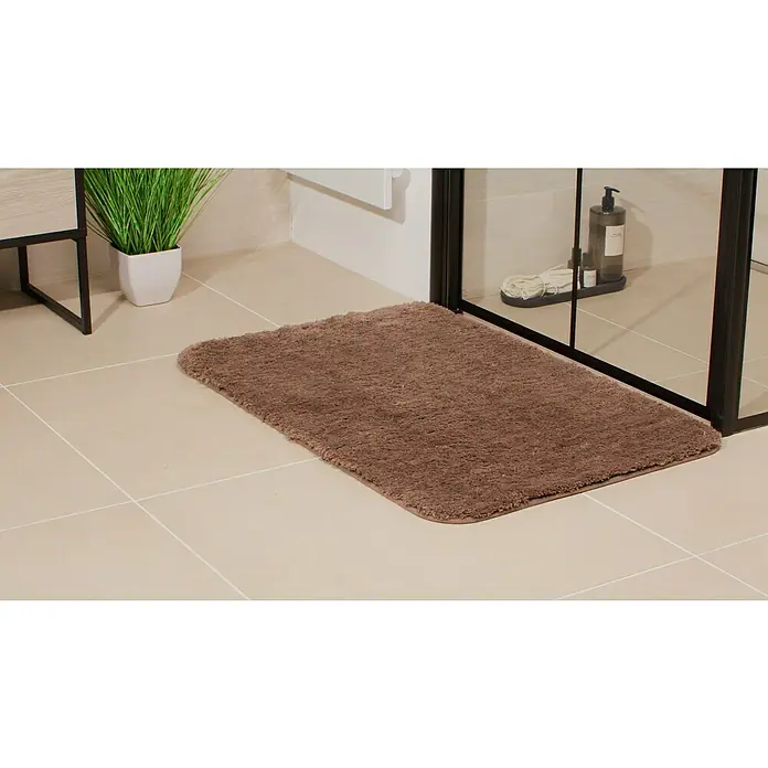 Camargue Badteppich Wuschel90 x 60 cm, Braun - Ein rechteckiger, brauner, flauschiger Badteppich liegt auf hellen Fliesen, neben einer Pflanze und einer Dusche.