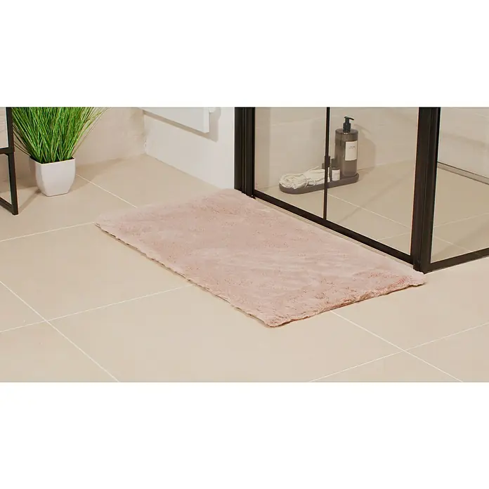 Badteppich Happy - 50 x 90 cm, Rosa, 100% Polyester - Flauschiger, rechteckiger, rosa Badezimmerteppich liegt vor einer Duschkabine auf beigen Fliesen. Eine Zimmerpflanze steht links.