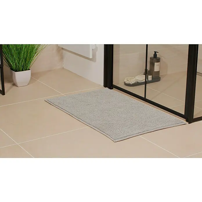 Camargue Badteppich Zottel - 50 x 80 cm, Manhattan, 100% Polyester - Graue Badematte vor einer Duschkabine auf hellbraunen Fliesen, die eine komfortable und stilvolle Ergänzung für jedes Badezimmer darstellt.