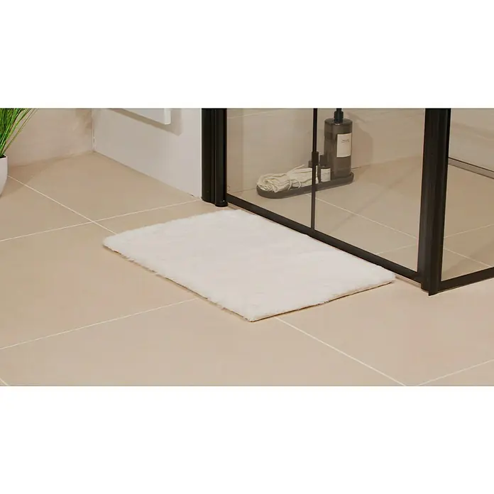 Badteppich Happy - 40 x 60 cm, Weiß, 100% Polyester - Ein weicher, rechteckiger, weißer Badezimmerteppich liegt vor einer modernen Duschkabine mit schwarzem Rahmen auf hellen Fliesen.