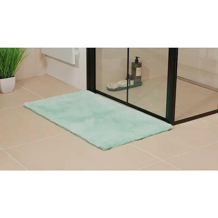 Badteppich Happy - 50 x 90 cm, Mint, 100% Polyester - Ein rechteckiger, hellgrüner, flauschiger Badteppich liegt vor einer Duschkabine auf hellen Fliesen. Zimmerpflanze und Pflegeprodukte sind im Hintergrund.