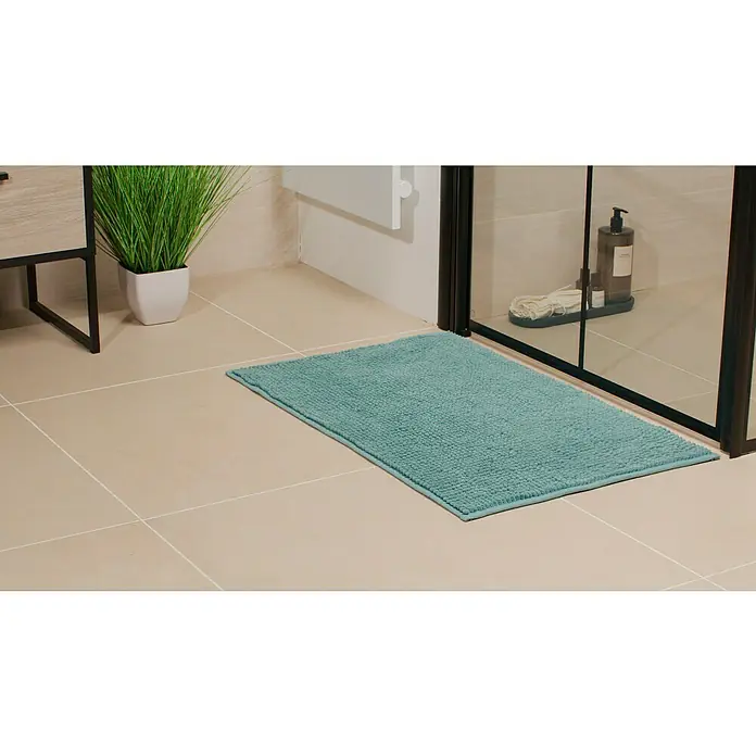 Camargue Badteppich Zottel - 50 x 80 cm, Petrol, 100% Polyester - Ein rechteckiger, türkisfarbener Badteppich liegt auf einem beigen Fliesenboden vor einer Duschkabine, wodurch er die Aufmerksamkeit auf sich zieht.