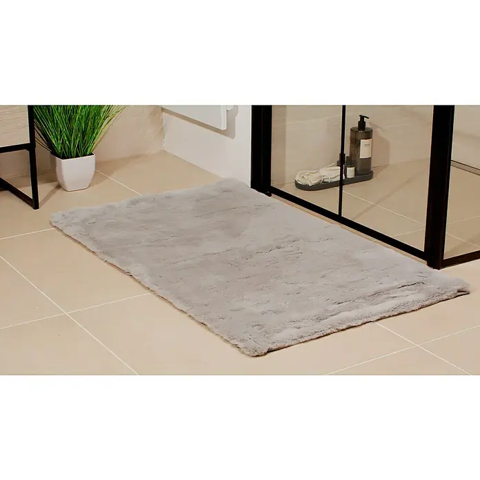 Badteppich Happy - 67 x 110 cm, Silber, 100% Polyester - Ein grauer, rechteckiger Badteppich aus weichem, flauschigem Material liegt auf hellbeigen Fliesen vor einer Duschkabine.