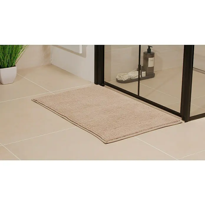 Camargue Badteppich Zottel50 x 80 cm, Light Grey, 100% Polyester - Ein rechteckiger, beiger Badvorleger liegt vor einer Duschkabine mit schwarzem Rahmen auf hellen Fliesen.