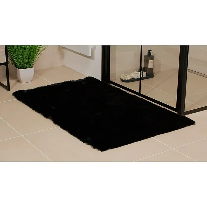 Badteppich Happy - 67 x 110 cm, Schwarz, 100% Polyester - Ein rechteckiger, schwarzer Badteppich aus Kunstfell liegt auf hellbeigen Fliesen vor einer Duschkabine mit schwarzen Rahmen.