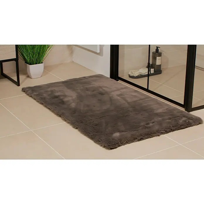 Badteppich Happy - 67 x 110 cm, Grau, 100% Polyester - Ein grauer, rechteckiger Badteppich aus weichem Stoff liegt auf hellen Fliesen vor einer Dusche und einer grünen Pflanze.