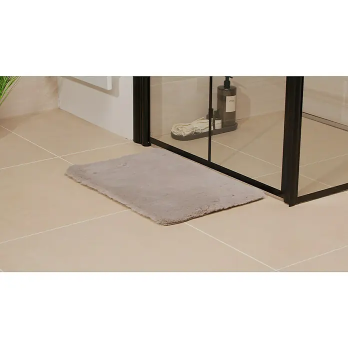 Badteppich Happy - 40 x 60 cm, Silber, 100% Polyester - Ein flauschiger, grauer Badteppich liegt vor einer modernen, schwarzen Duschkabine auf hellen Fliesen.