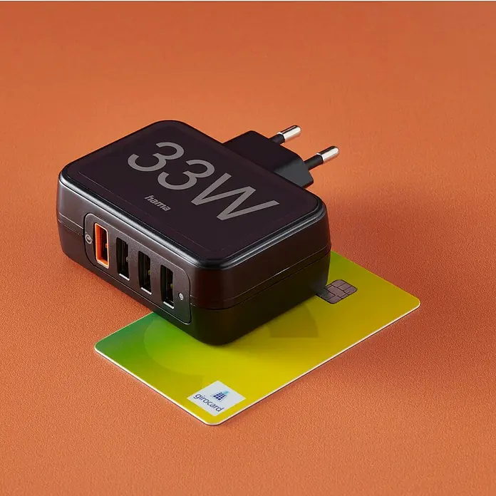 Hama USB-Adapter Schnellladegerät 4x USB-A 33W - Schwarz, USB A-Stecker - Produktansicht des Hama 33W USB-Ladegeräts mit vier Anschlüssen, das auf einer Girokarte platziert ist, vor orangefarbenem Hintergrund.