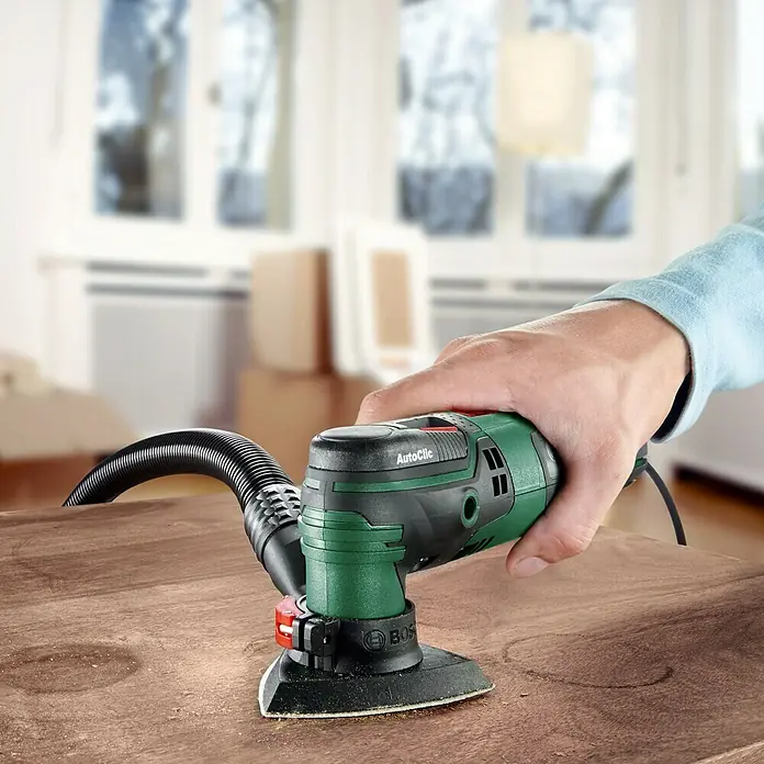 Bosch Multiherramienta PMF 250 CES - 250 W, Sistema STARLOCK - Primer plano de una lijadora delta Bosch en acción, utilizándose sobre una superficie de madera. La herramienta es verde y negra, con aspiración de polvo.
